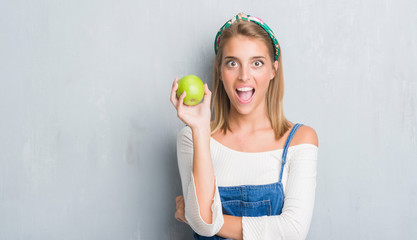 Beautiful young woman over grunge grey wall eating green apple scared in shock with a surprise face, afraid and excited with fear expression