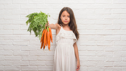 Poster - Young hispanic kid over white brick wall holding fresh carrots with a confident expression on smart face thinking serious