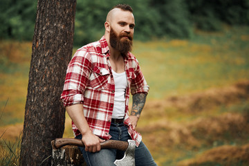 Wall Mural - brutal lumberjack in red shirt with beard holding axe sitting in Forest after work