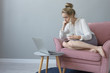 Indoor shot of attractive young female with hair bun sitting barefooted in armchair with cup of coffee and watching webinar via laptop computer, learning online, having focused concentrated look