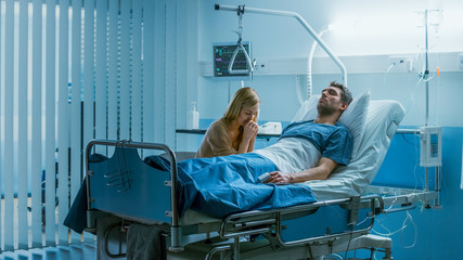 Wall Mural - In the Hospital Sick Man Lying on the Bed, His Visiting Wife Sorrowfully Sits Beside Him and Prays for His Rapid Recovery. Blue and Melancholy Color of the Scene.