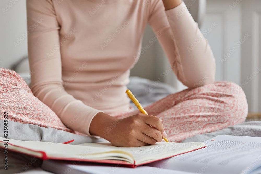 Unrecognizable woman dressed in casual nightwear, writes in notebook, has inspiration for studying. Girl makes notes in diary what happend with her during day before sleeping. Focus on hand with pen - obrazy, fototapety, plakaty 