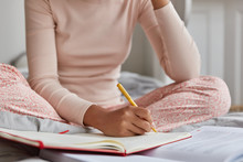 Unrecognizable Woman Dressed In Casual Nightwear, Writes In Notebook, Has Inspiration For Studying. Girl Makes Notes In Diary What Happend With Her During Day Before Sleeping. Focus On Hand With Pen