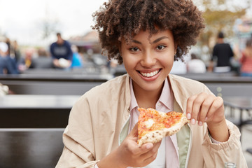 Photo of satisfied teenage girl with dark healthy skin, enjoys delicious meal, holds piece of pizza, has snack dressed in jacket, spends free time in outdoor pizzeria or cafe, has toothy smile.