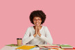 Positive young African American girl concentrated on course work, writes ideas, keeps hands together and smiles positively, happy to solve problems with project, poses against pink background