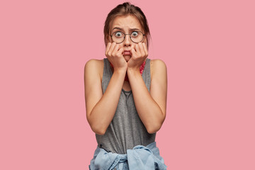 Wall Mural - Terrified trembling woman stares in stupor with eyes popped out, expresses fear and shock, wears casual outfit and round spectacles, poses against pink background. Nervous girl poses indoor.