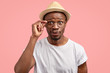 Photo of curious African American male looks with mysterious expression, keeps hand on rim of spectacles, wears casual clothes, pouts lips, listens something with interest, poses over pink background
