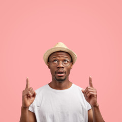 Indoor shot of stupefied dark skinned young man in fashionable hat casual white t shirt, poses against pink background with copy space, shows something above. Ethnicity and advertisement concept