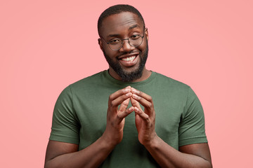 Wall Mural - Horizontal shot of satisfied businessman keeps hands together, looks curiously, has pleasant talk with interlocutor, has dark thick beard, dressed in casual t shirt, poses against pink background