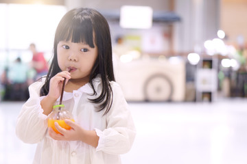 Asian child cute or kid girl long black hair with white clothes stand and drink orange juice by tube or straw for refreshing and vitamin C in the department store with sunlight