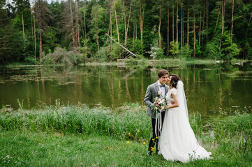 Wall Mural - Portrait of an attractive groom and bride on nature in the park. Happy and joyful moment. Romantic couple of newlyweds kissing near pond. Wedding ceremony near lake.