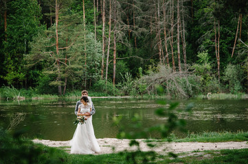 Wall Mural - Portrait of an attractive groom and bride standing on nature in the park. Happy and joyful moment. Romantic couple of newlyweds near pond. Wedding ceremony near lake. Photography. Photo.