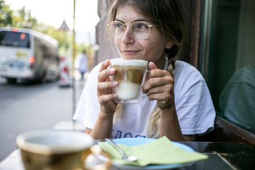 Wall Mural - Cute millennial young pretty woman with braids relaxed and chill sits in cafe terrace outside, enjoys coffee drink during summer lunch break, drinks latte layered cappuccino from glass cup