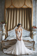 Beautiful young bride, sexy brunette model girl in the stylish and fashionable wedding dress with naked shoulders sits on the bed and posing in luxury vintage interior