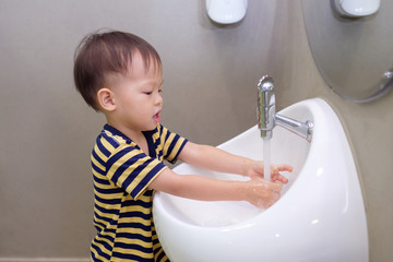 Wall Mural - Cute little Asian 2 years old toddler baby boy child washing hands by himself on white sink and water drop from faucet in public toilet / bathroom for kids, Sanitation / hygiene concept