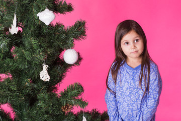 Children, holidays and christmas concept - little girl near christmas tree on pink background