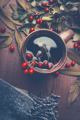 Wall Mural - Autumn concept. Cup of herbal tea with red fall berries decorated with leaves on dark rustic wooden table background , top view, still life