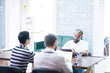© Seventyfour - Positive handsome young mixed race manager with beard wearing glasses sitting at table with colleagues in loft board room and analyzing statistics presented by staff