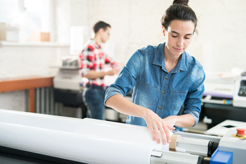 Serious confident young woman  with hair bun loading wide format printer while working at printing house