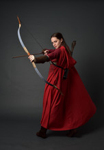 Full Length Portrait Of Brunette Girl Wearing Red Medieval Costume And Cloak, Holding A Bow And Arrow. Standing Pose On Grey Studio Background.