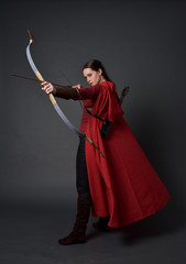 Canvas Print - full length portrait of brunette girl wearing red medieval costume and cloak, holding a bow and arrow. standing pose on grey studio background.