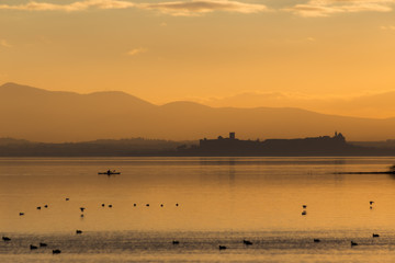 Beautiful view of Trasimeno lake (Umbria, Italy) at sunset, with orange tones, birds on water, a man on a canoe and Castiglione del Lago town on the background