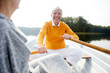 Cheerful excited handsome elderly man in shirt and yellow sweater sitting on boat and rowing with oars while talking to woman