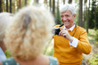 Jolly excited handsome senior man in yellow sweater photographing friends on camera and laughing while asking them to pose in forest