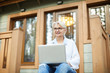 © pressmaster - Smiling modern working senior lady in glasses sitting on porch of cottage and looking into distance while working on project using laptop
