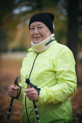 Wall Mural - An elderly woman in a green down jacket is standing in a park with sticks for nordic walking