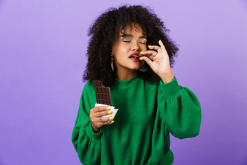 Poster - Young pretty african woman isolated over violet background eat chocolate.