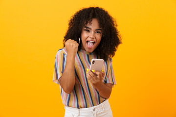 Sticker - Emotional african woman posing isolated over yellow background using mobile phone make winner gesture.