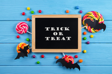 Wall Mural - Trick or treat text written on blackboard with decorative paper bats, eyeball shaped candy, gummy worms, spiders & bones. Halloween decor concept. Background, copy space, close up, top view, flat lay.