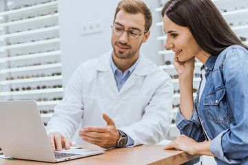 smiling woman standing near male oculist while he pointing by hand at laptop screen in optics