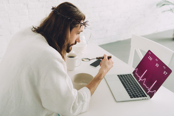 Wall Mural - concentrated Jesus eating corn flakes on breakfast at table with laptop with graph on screen in kitchen at home