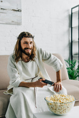 Canvas Print - smiling Jesus in crown of thorns watching tv and sitting with bowl of popcorn on sofa at home