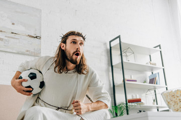 Wall Mural - emotional Jesus in crown of thorns sitting on sofa with soccer ball and watching football match at home