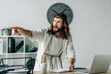 Aggressive Jesus In Crown Of Thorns And Robe Writing In Textbook And Pointing By Finger At Table With Laptop In Modern Office