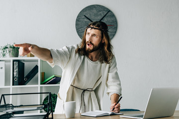 Wall Mural - aggressive Jesus in crown of thorns and robe writing in textbook and pointing by finger at table with laptop in modern office