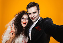 Photo Of Beautiful Zombie Couple Bridegroom And Bride Wearing Wedding Outfit And Halloween Makeup Laughing While Taking Selfie, Isolated Over Yellow Background