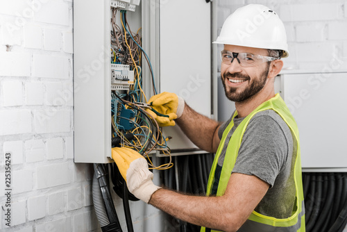 Fotomural smiling handsome electrician repairing electrical box with pliers in corridor an