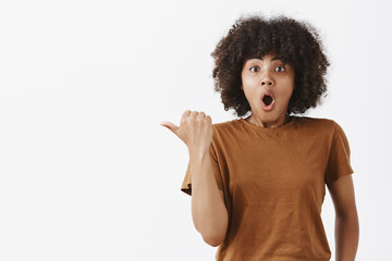 Wall Mural - uplifted excited cute dark-skinned girl with curly hair in brown trendy t-shirt pointing left with t