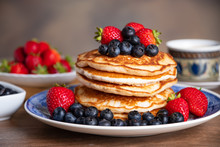Stack of pancakes with blueberries and strawberries on a blue and white plate with bowls of strawberries and blueberries and a cup of coffee in the background. 