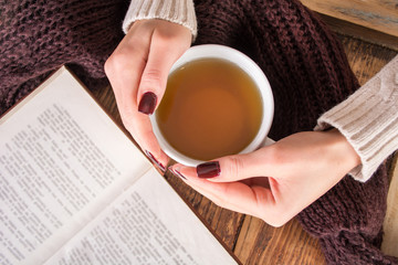 Girl hands with cup of tea and open book on wooden desk. Woman wears wool sweater. Retro and vintage concept. Top view, selective focus and close up