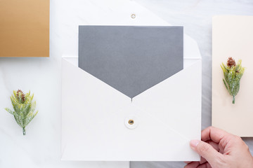 Wall Mural - Top view of hand holding blank grey card in white envelop with pine leaf on two layer step of marble table top.Mock up template for display of design.modern business items.