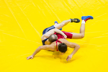 Two young men in blue and red wrestling on a yellow wrestling carpet in the gym