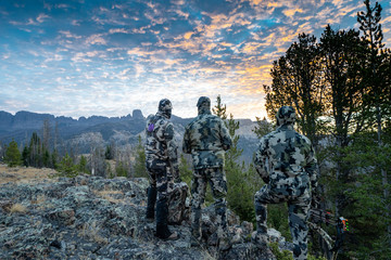 three adult male hunter friends, unrecognizable, stand on a mountain ridge looking for elk to hunt d
