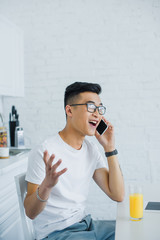 Wall Mural - emotional young asian man talking by smartphone while sitting in kitchen