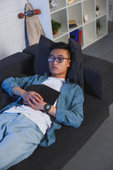 Wall Mural - high angle view of young man in eyeglasses holding book and sleeping on couch