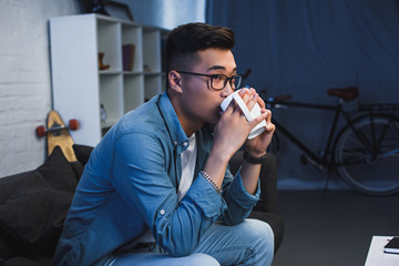 Wall Mural - young asian man in eyeglasses sitting on couch and drinking from cup